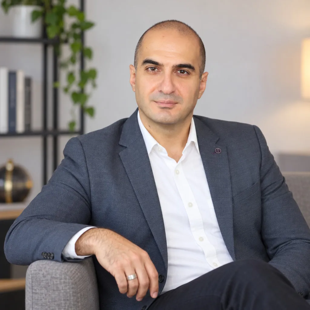 Rem Darbinyan in a grey blazer seated in an armchair, arm resting on the chair, bookshelf behind